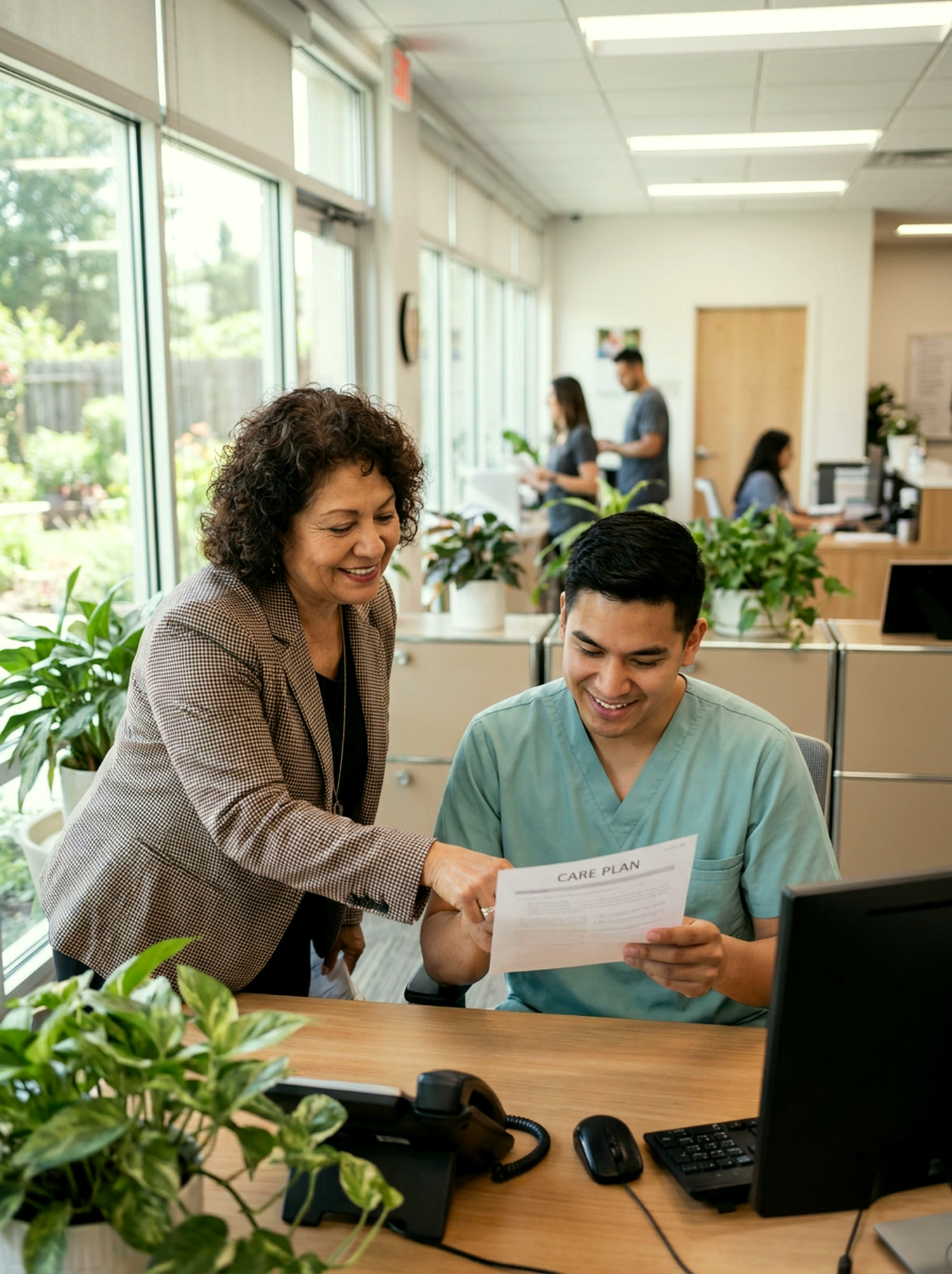 An experienced case manager mentors a newer colleague in teal scrubs as they review a care plan together at a sunlit office table.